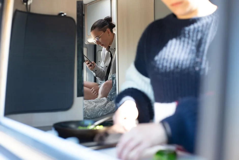 Person kocht mit einer Pfanne im Innenraum eines Campervans, im Hintergrund zwei weitere Personen