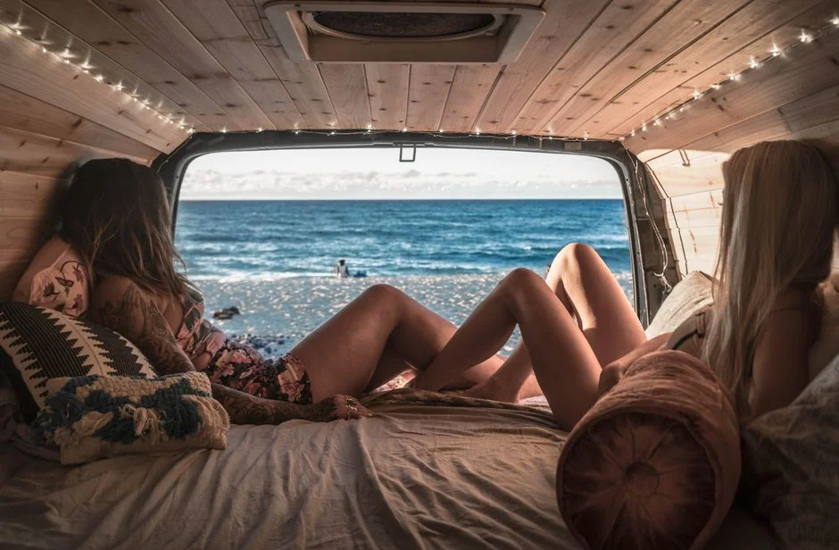 Zwei Personen liegen in einem Van mit Holzverkleidung an Decke und Seitenwand, Blick aufs Meer