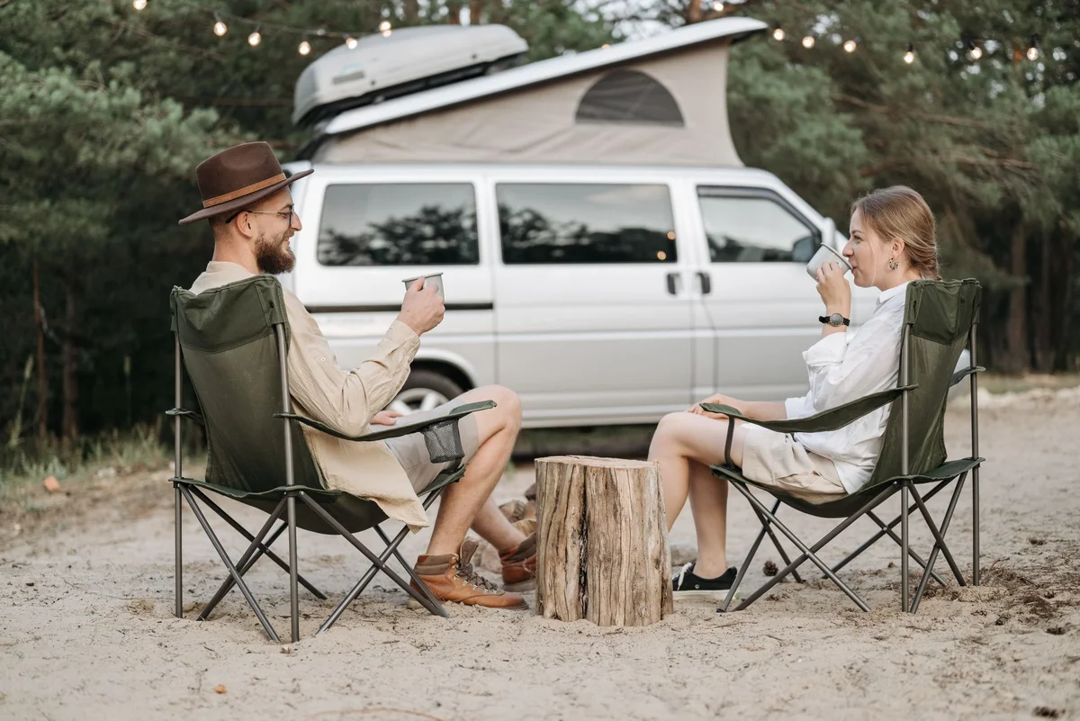 Paar sitzt vor einem Camper mit Aufstelldach im Wald und trinkt Kaffee
